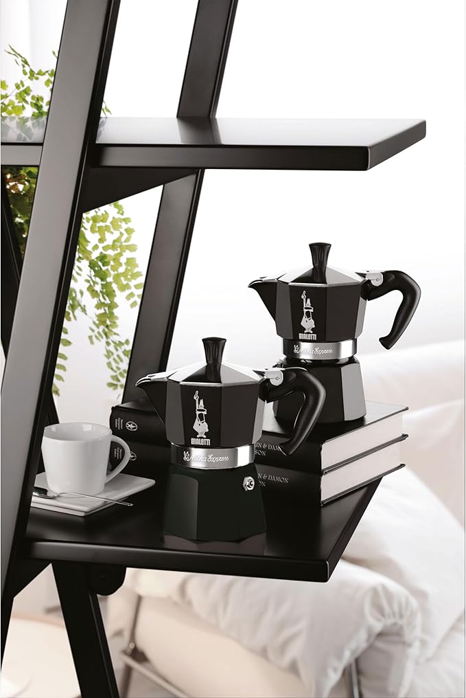 cafetera italiana Bialetti Moka express negra de aluminio con mango ergonómico y válvula de seguridad en estante negro junto a taza y libros