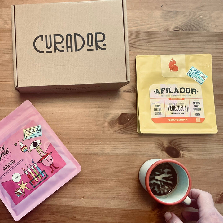 Caja de suscripción Curador con café en grano y molido de especialidad junto a una taza de café servido en mano sobre mesa de madera
