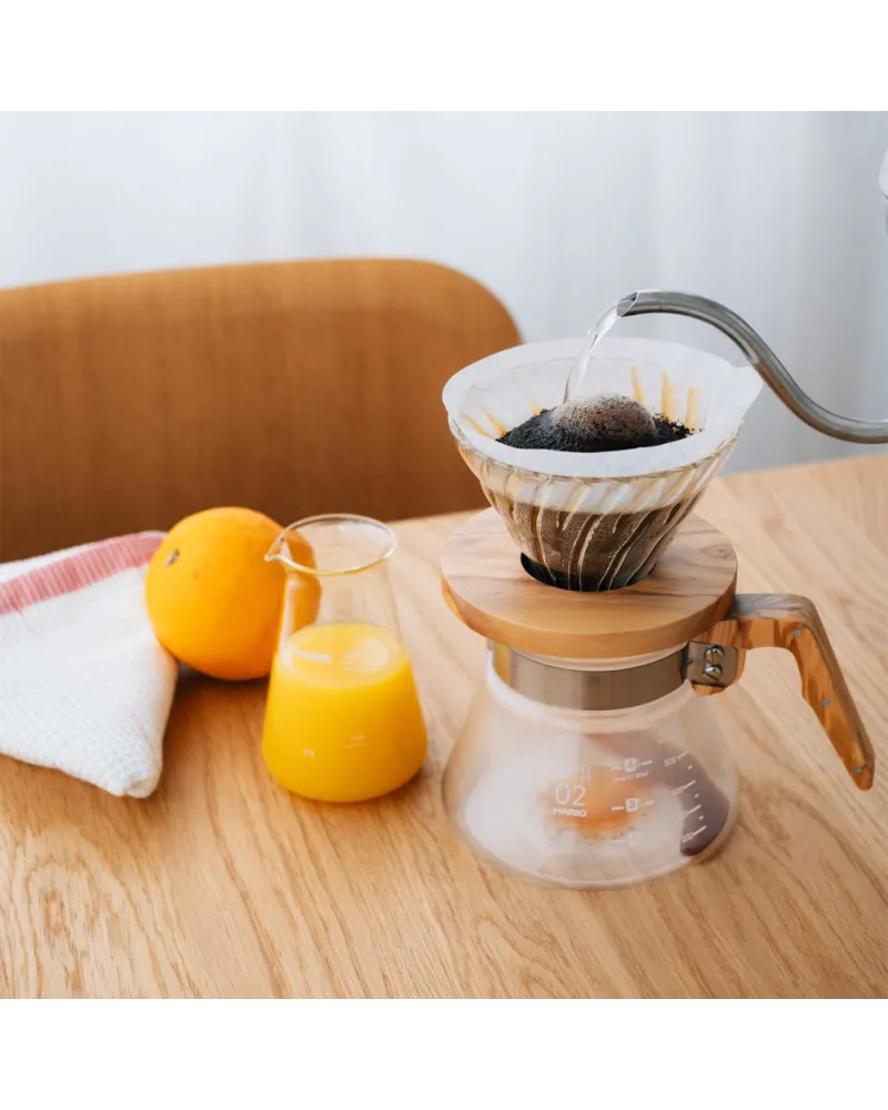 cafetera V60 dripper de vidrio con base de madera preparando café filtrado sobre mesa de madera con jarra de jugo de naranja y naranja fresca al lado