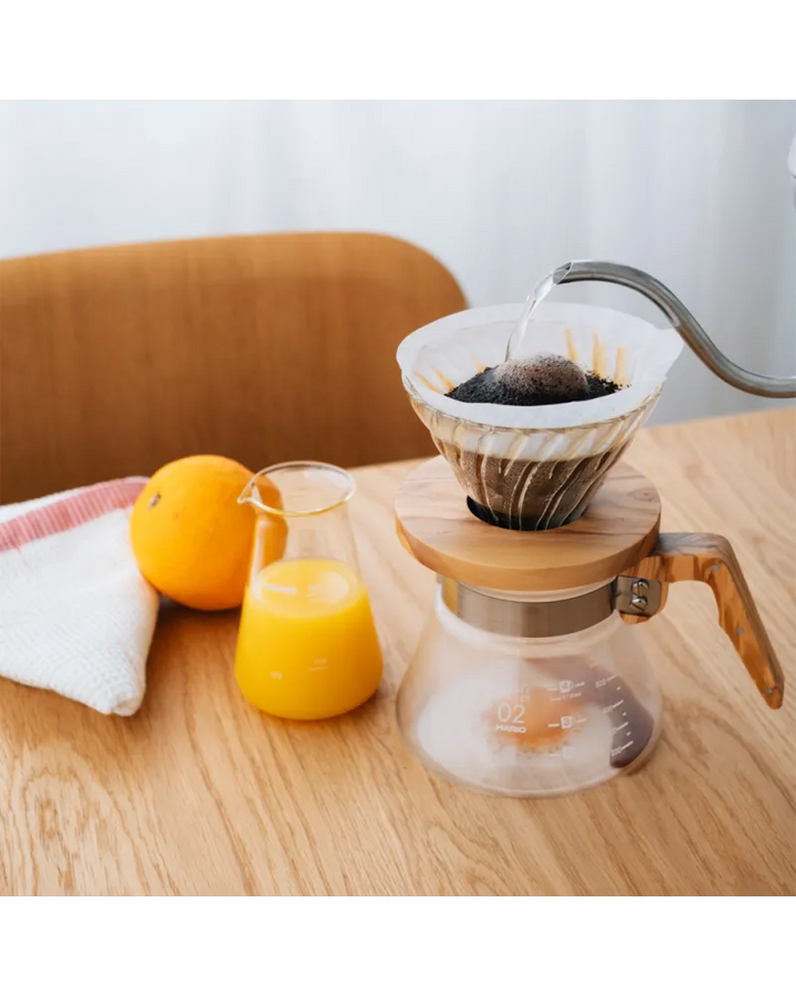 cafetera V60 dripper de vidrio con base de madera preparando café filtrado sobre mesa de madera con jarra de jugo de naranja y naranja fresca al lado